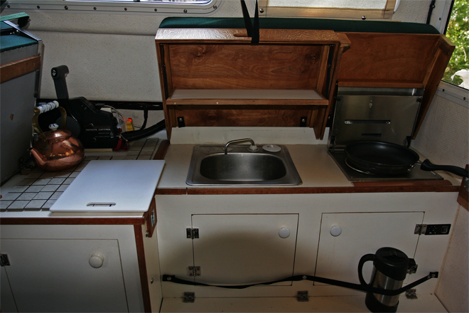 Covers are raised and helm seat pivots up to use galley. Hot items can be placed on ceramic tiles under helm seat. Small drop-down shelf inside forward cover is made of starboard trimmed in teak. Snap on elastic bands are handy for holding essentials (spelled COFFEE press) when underway.
